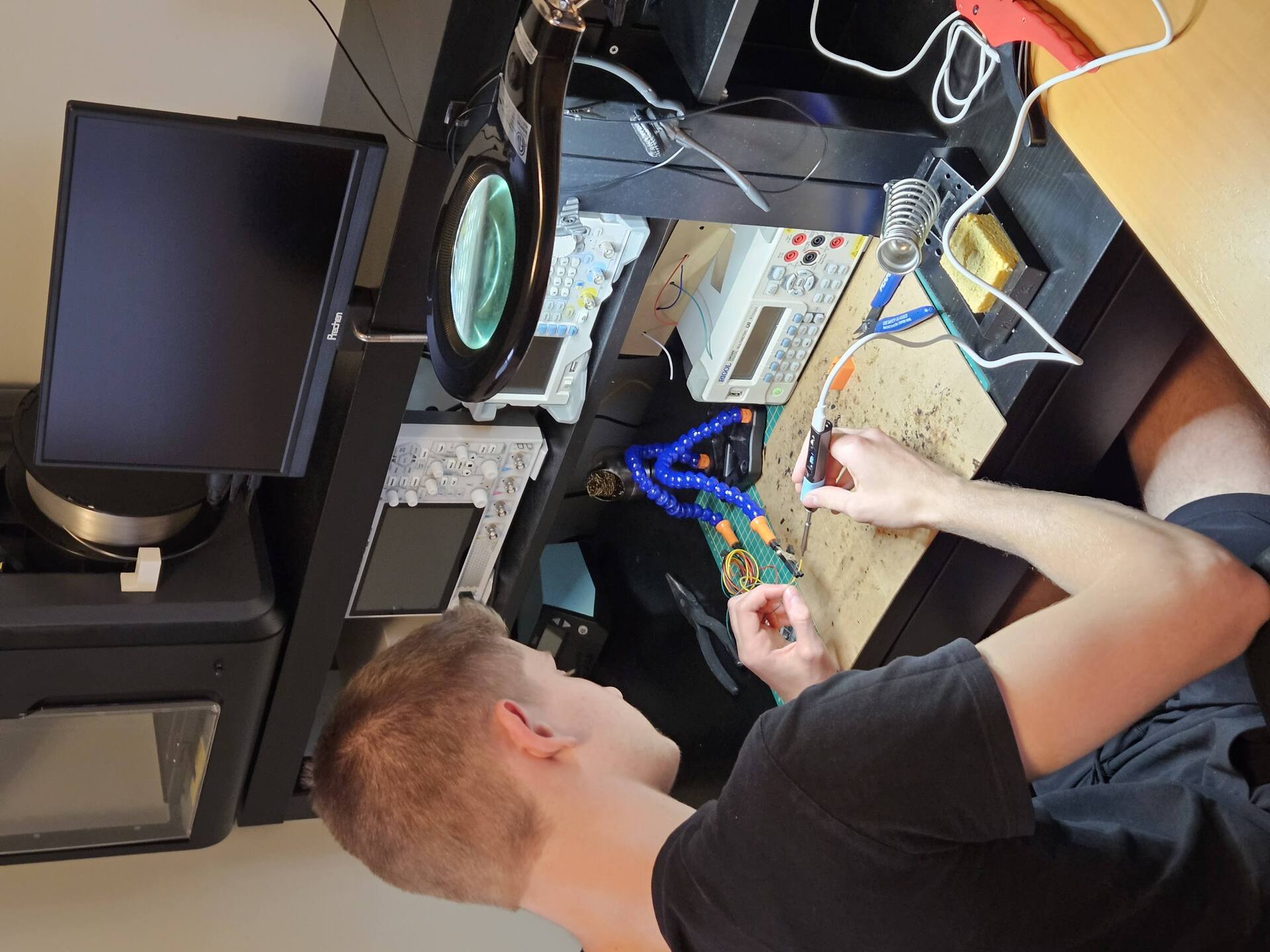 Anthony Nistor soldering at an electronics bench, with a 3D printer and test instruments behind him.